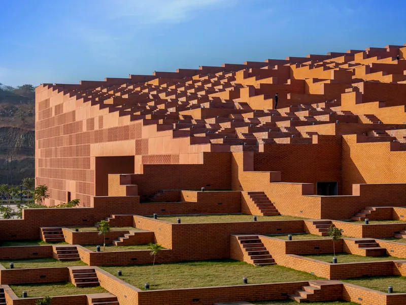 Cascading brick facade with pixelated massing of orange and brown blocks beneath a clear blue sky