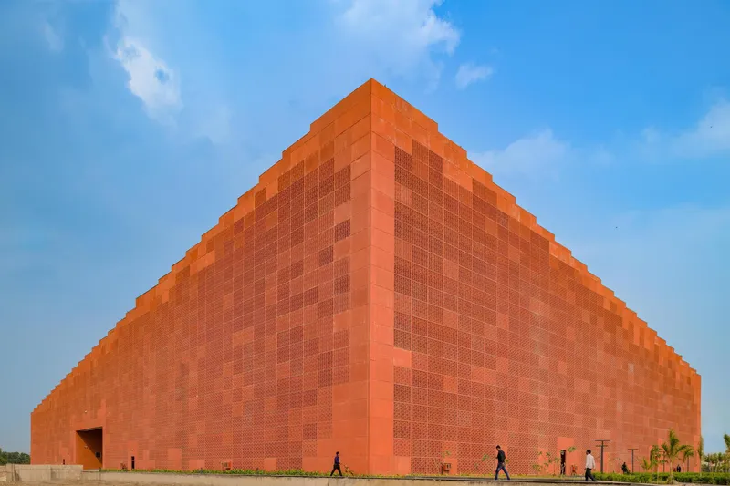Pyramidal orange brick volume with textured screen panels and figures walking along the base at dusk