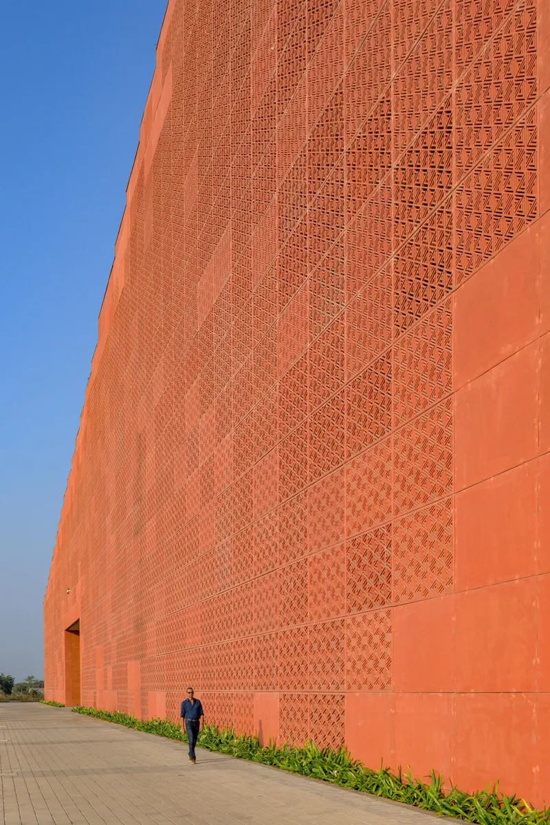 Close-up of orange brick wall with patterned perforations and a figure walking along the paved edge