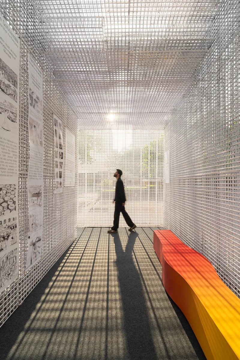Gallery corridor with perforated white mesh walls and ceiling as figure walks past gradient orange bench