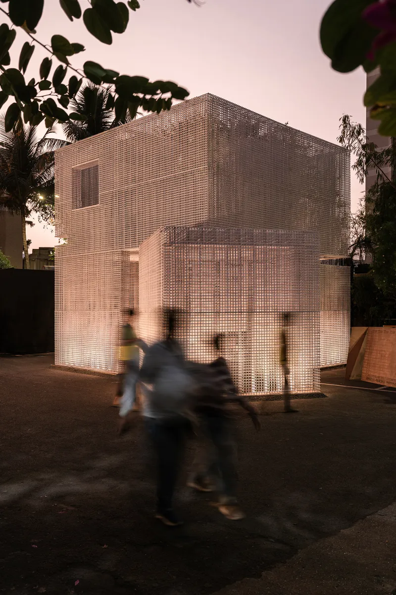 Two-story pavilion wrapped in translucent perforated screens with a motorbike passing at dusk