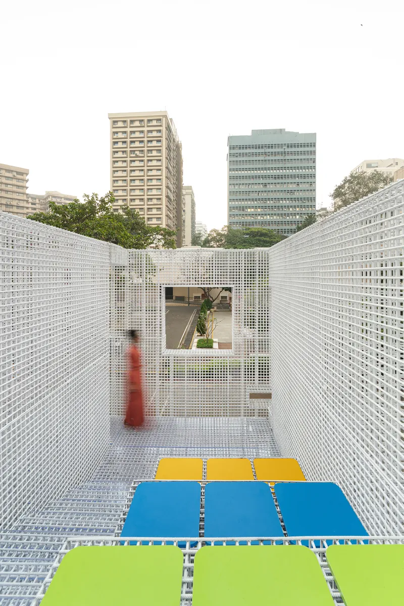 White perforated metal screen corridor with colored floor panels and a visitor in red framed by urban towers