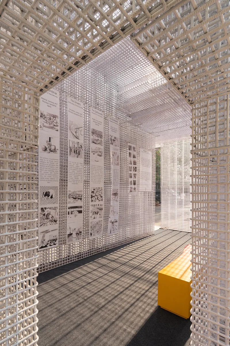 View through white perforated screen showing exhibition graphics on walls and a yellow bench on dark flooring