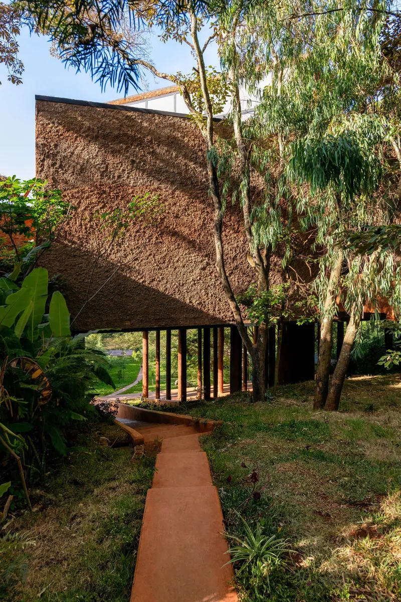Woven thatch roof pavilion elevated on timber columns with a red earth pathway leading through mature trees
