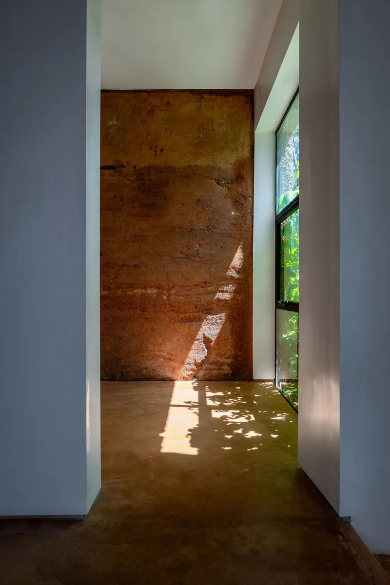 Corridor with weathered rammed earth feature wall lit by sidelights casting leaf shadows on concrete floor