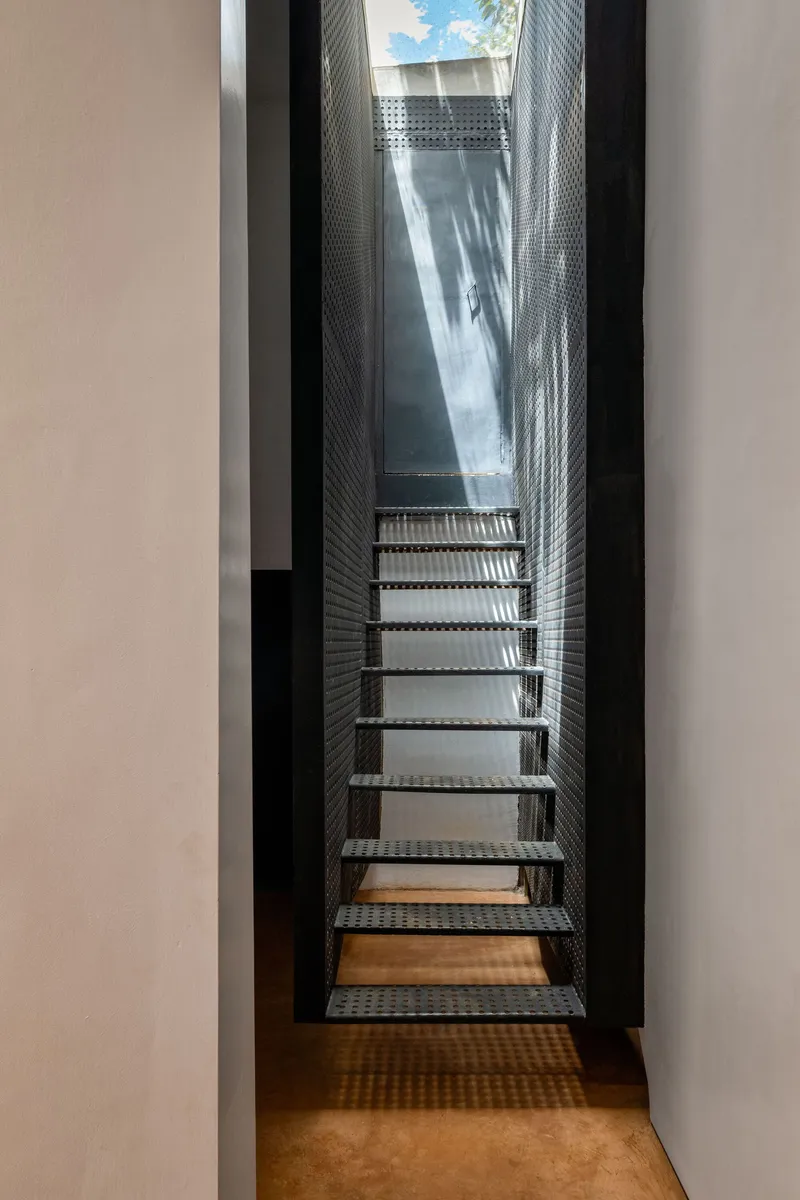 Perforated metal staircase rising through narrow lightwell toward skylight opening above