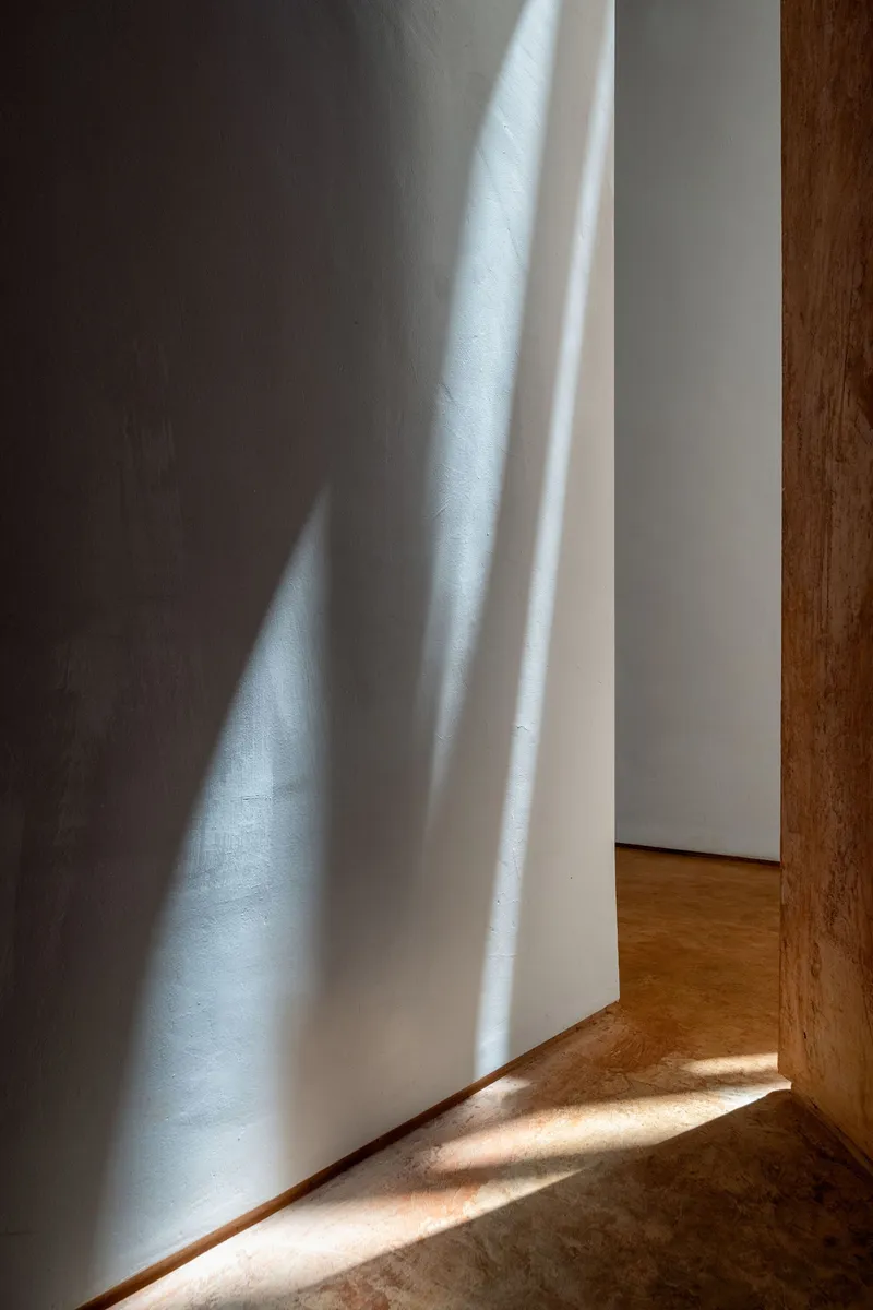 Interior corner where white plaster walls meet timber-edged flooring with angled morning light