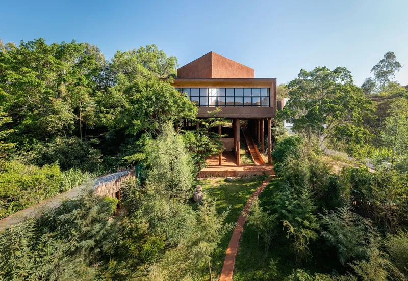 Elevated red earth structure on timber supports surrounded by terraced garden beds and native trees