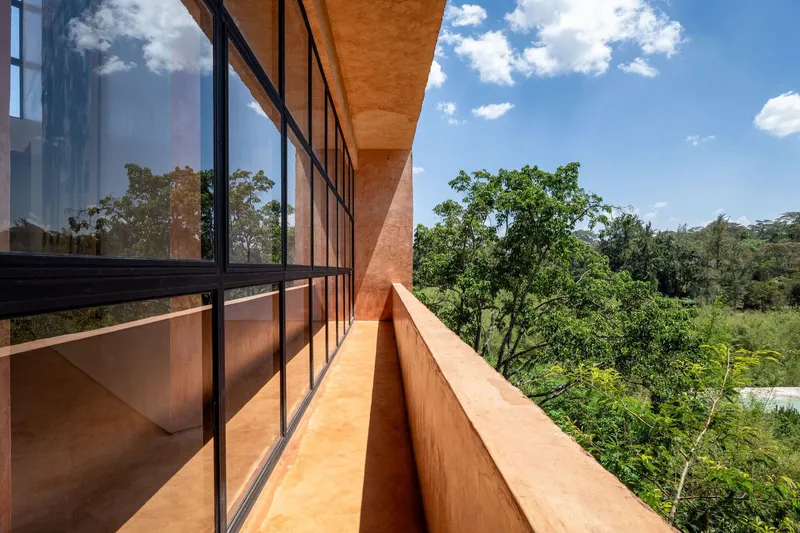 Exterior balcony with red earth parapet wall and steel-framed glazing overlooking green canopy