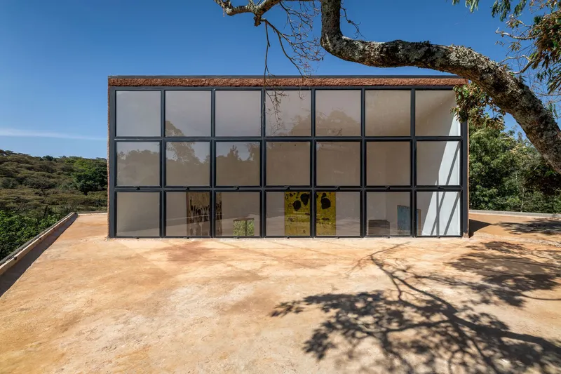 Grid facade of steel-framed translucent panels facing a red earth terrace under tree shade