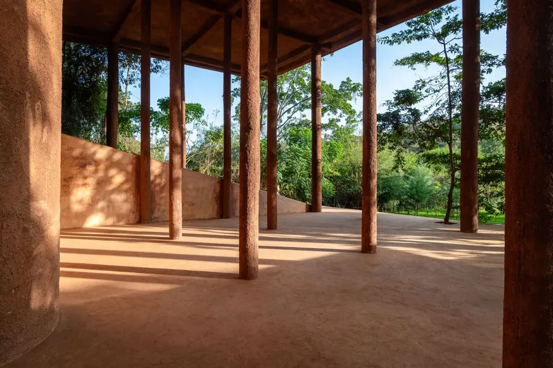 Open-air pavilion with timber columns and exposed truss ceiling casting dappled shadows on floor