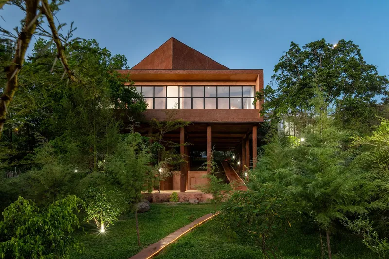 Facade with pyramidal red earth roof and raised pavilion emerging from landscaped hillside at dusk
