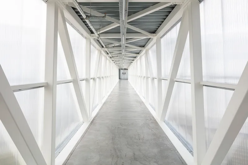 Enclosed corridor with white diagonal steel bracing and translucent polycarbonate cladding under an overcast sky