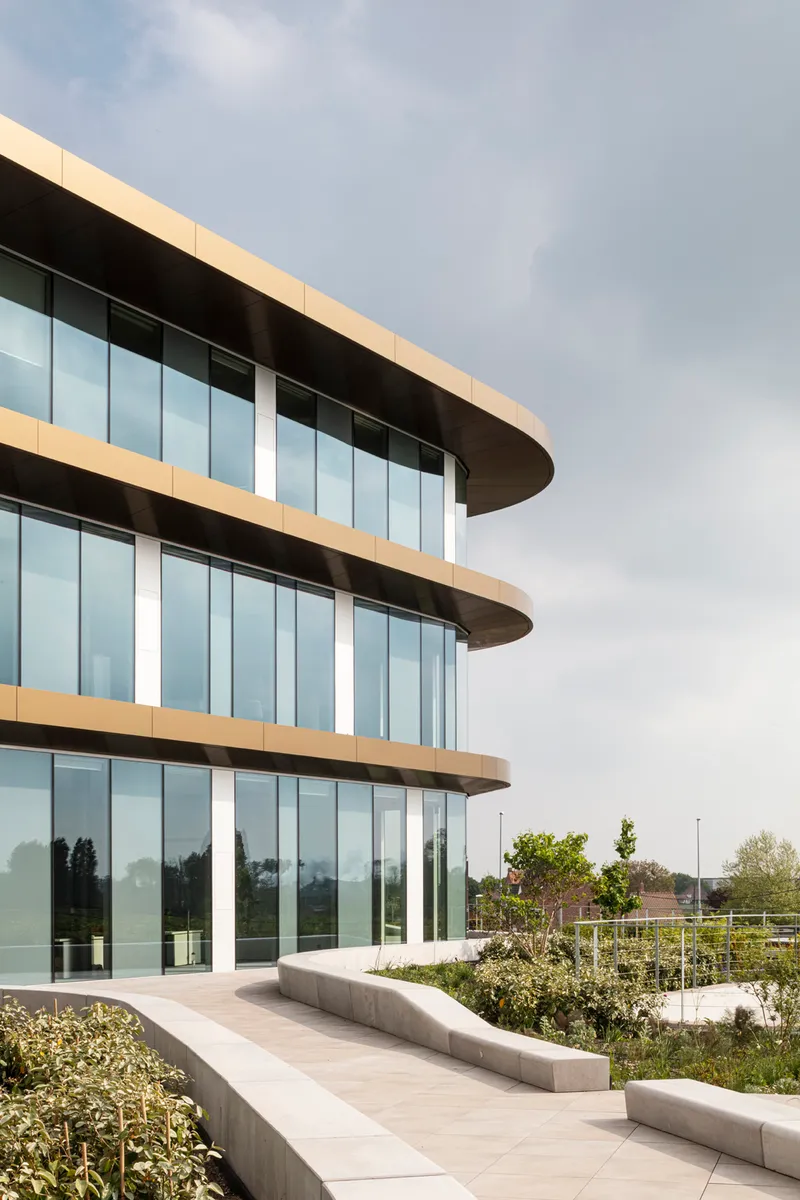 Exterior terrace with curved glass facade, stone pavers and planted beds under an overcast sky