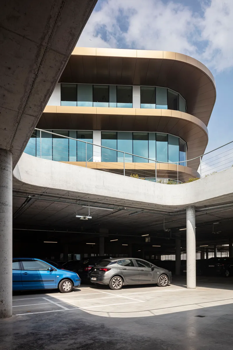 Rounded glass facade rising above an exposed concrete parking structure with parked cars