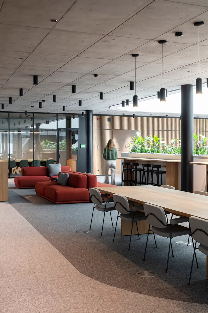 Open workspace with concrete ceiling, pendant lights, and a figure passing the communal dining table