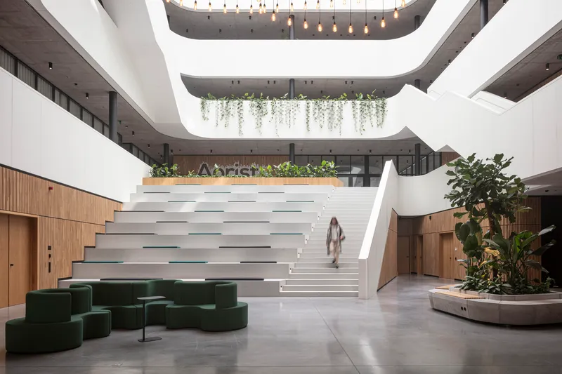Multi-storey atrium with a wide white staircase, hanging plants on balconies, and green modular seating below