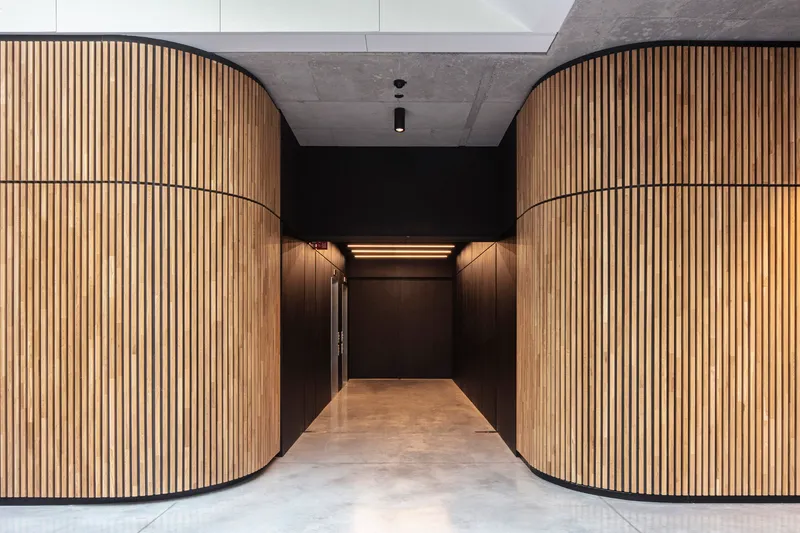 Corridor framed by curved timber slat walls leading to a dark-paneled elevator lobby under concrete ceiling