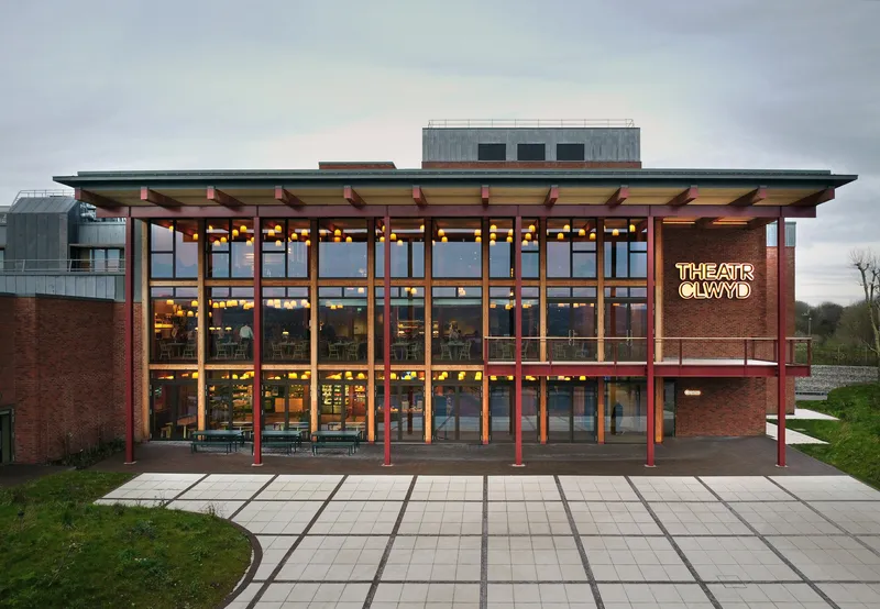 Glazed entrance facade with timber frame columns illuminated at dusk beside paved plaza