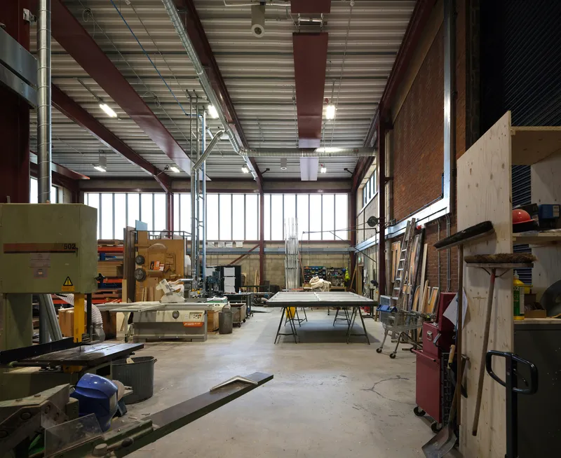 Workshop interior with exposed steel beams, corrugated metal ceiling, and worktables surrounded by tools and materials
