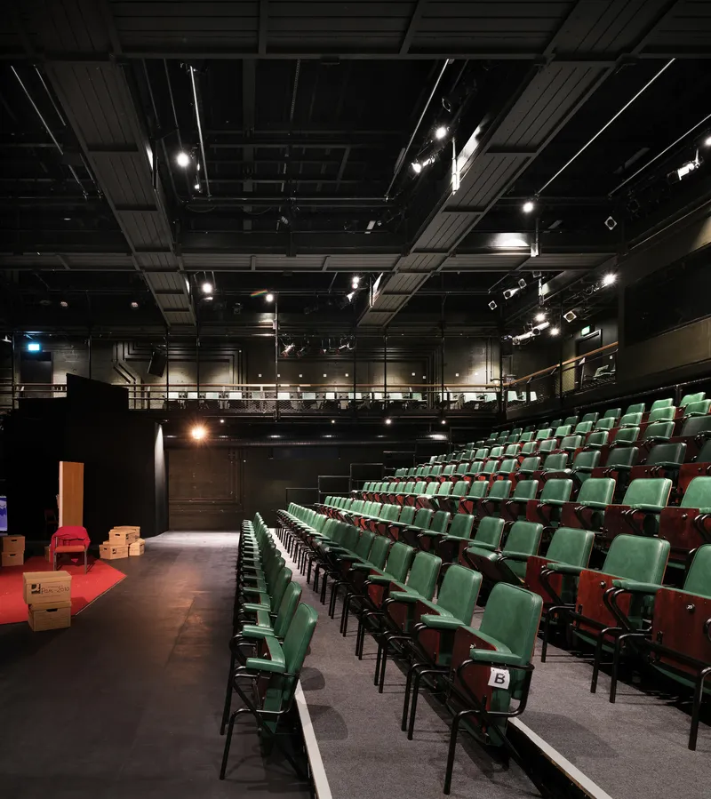 Theater auditorium with raked seating, exposed black ceiling with stage lights, and view toward the stage