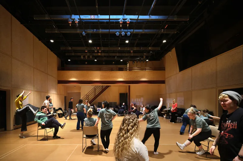 Rehearsal room with plywood-paneled walls, exposed technical ceiling, and students stretching in a circle