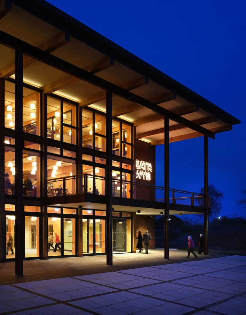 Three-story glass facade with timber ceiling and steel columns lit warmly at blue hour