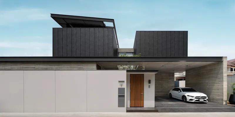 Street facade with pale garage doors, timber entry and dark metal volumes above board-formed concrete carport