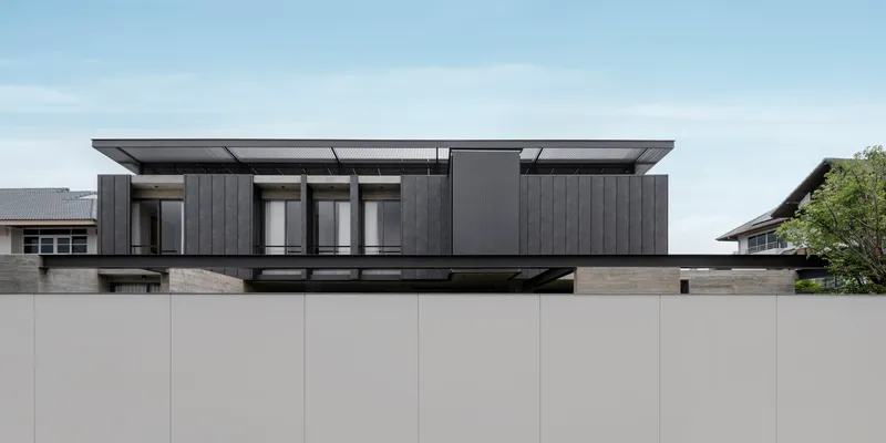 Street view of the dark timber-clad upper volume floating above a beige boundary wall