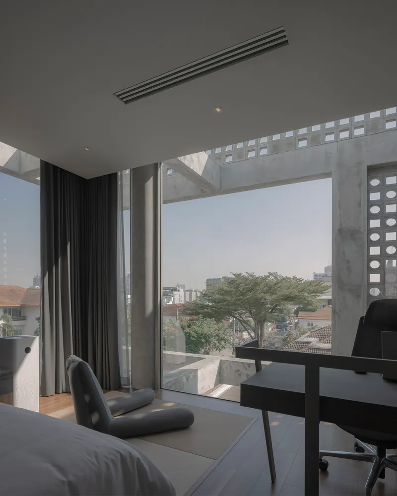 Bedroom window framed by perforated concrete beams overlooking neighborhood rooftops and a tree