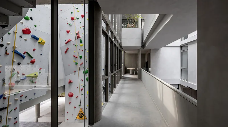 Concrete circulation corridor with colorful climbing wall visible through glass partition and planted courtyard beyond
