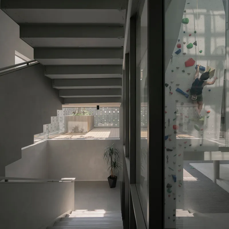 Concrete staircase ascending past a glazed climbing wall with a person scaling the holds in daylight