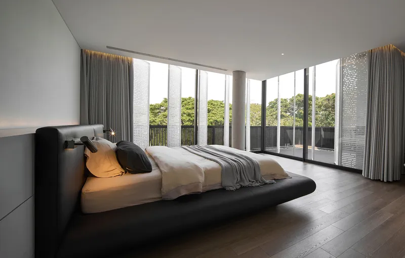 Corner bedroom with floor-to-ceiling glass walls overlooking greenery and grey curtains filtering daylight