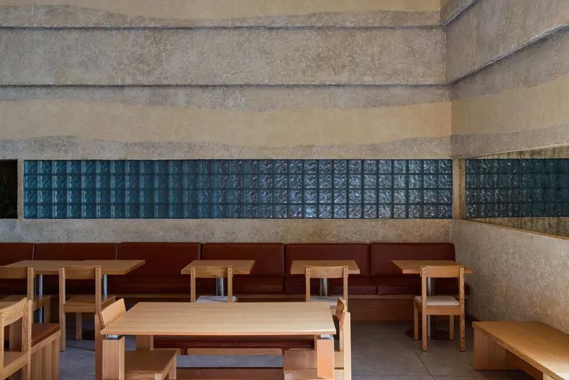 Banquette seating along layered concrete wall with horizontal band of blue glass block and timber tables