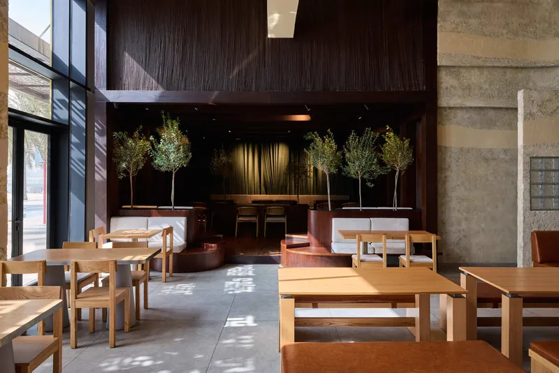 Dining hall with timber tables facing a stage alcove with olive trees in afternoon sunlight