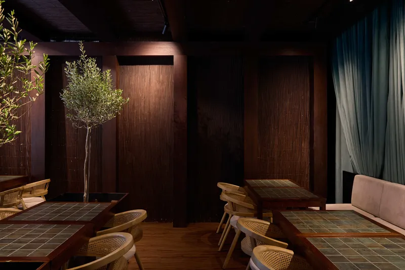 Wood-paneled wall with vertical slats between planted olive trees and tiled dining tables under warm overhead lighting