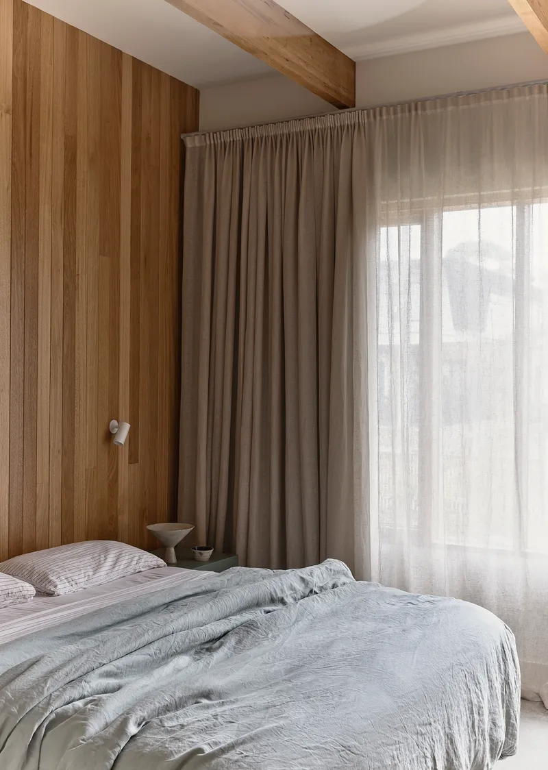 Bedroom with timber paneling and exposed beam ceiling beside layered linen curtains in soft light