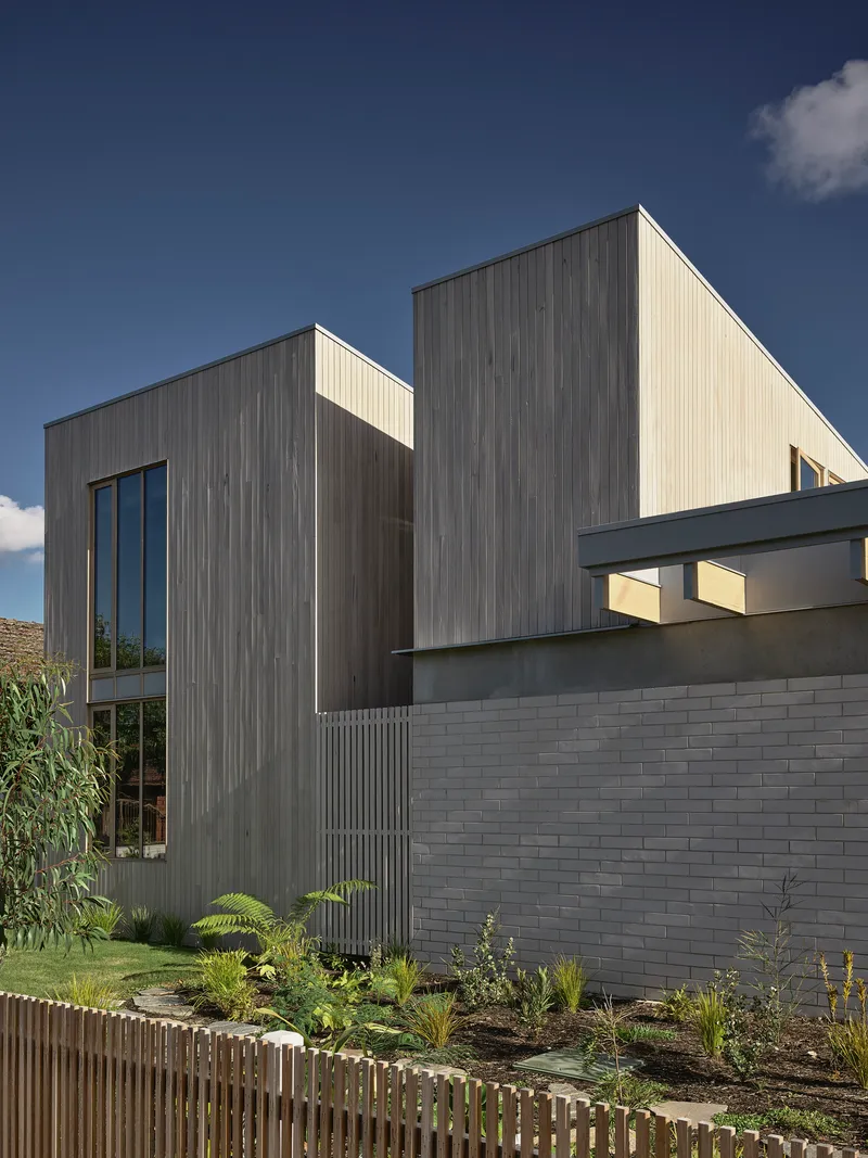 Street facade showing vertical timber cladding above a grey brick base with planted garden bed and timber fence