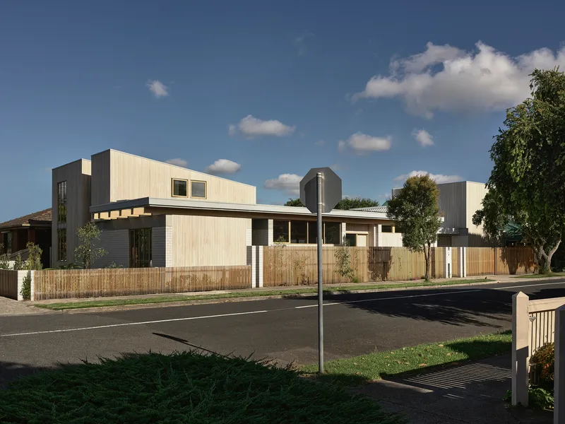 Street view of the residence with vertical timber cladding, timber fence, and mature street trees under scattered clouds