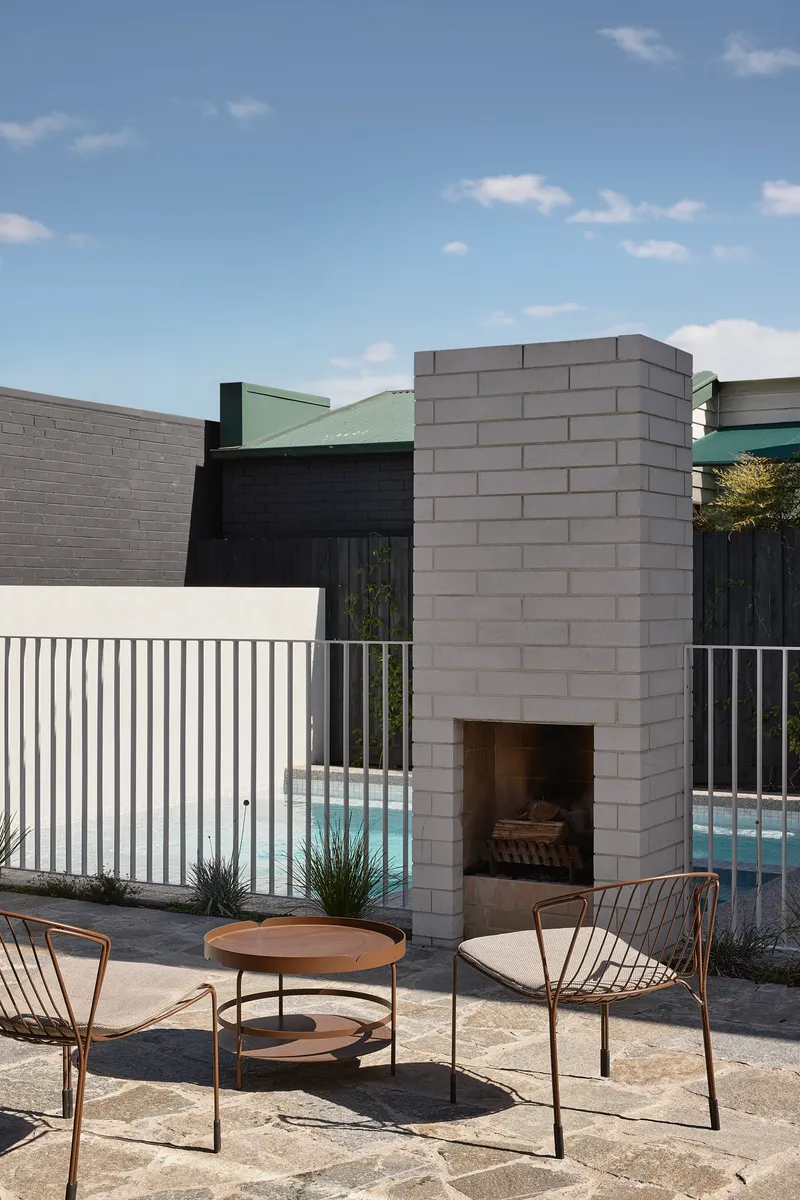 Outdoor concrete block fireplace with wire mesh chairs and round timber table beside the pool