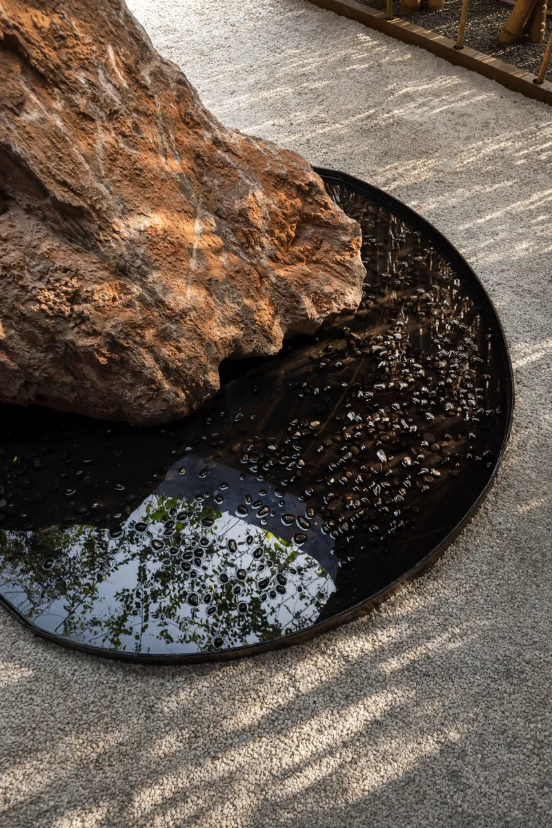 Close-up of weathered boulder resting on circular black reflecting pool with gravel floor casting rippled reflections