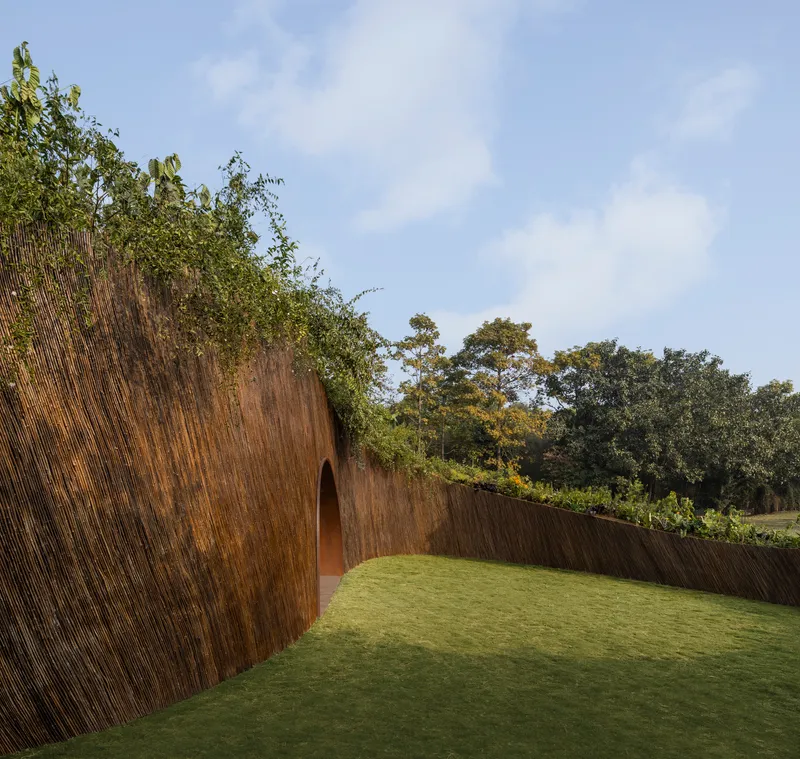 Exterior view of curved bamboo wall with arched passage enclosing lawn courtyard under partly cloudy sky