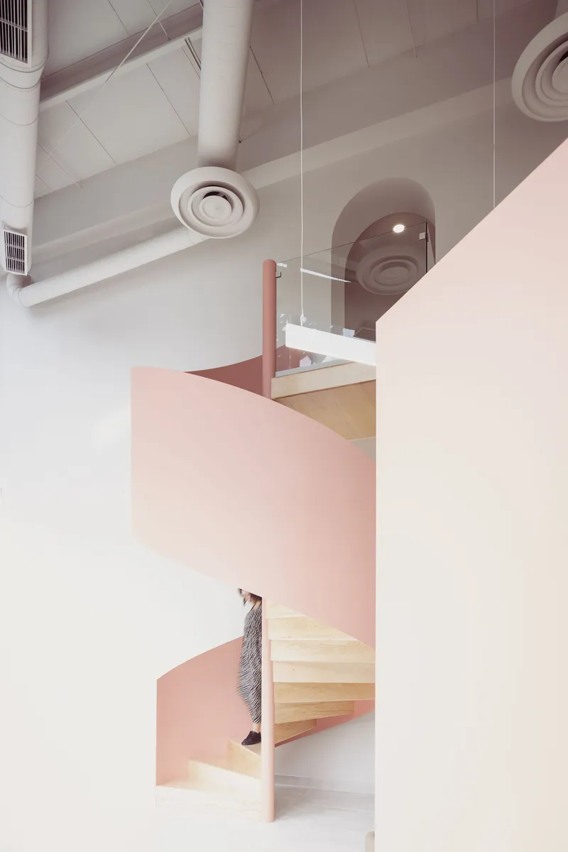 Spiral staircase with light wood treads and pink partition walls beneath exposed ductwork and ceiling vents