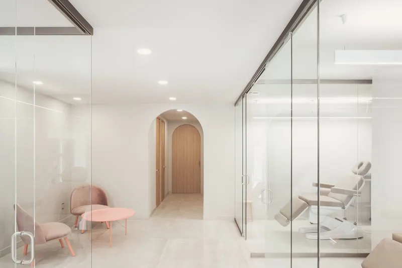 Corridor with floor-to-ceiling glass partitions revealing treatment rooms and a timber-clad arched doorway beyond