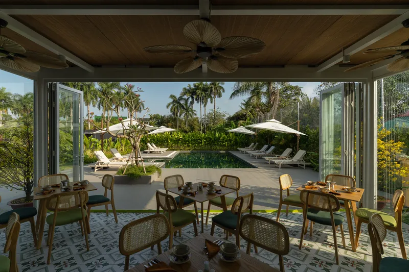 Open-air dining room with folding glass walls overlooking swimming pool surrounded by palm trees and loungers