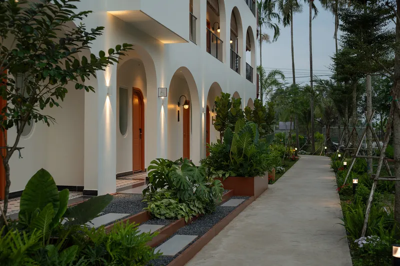 White arched facade with planted beds and concrete walkway at dusk under palm canopy