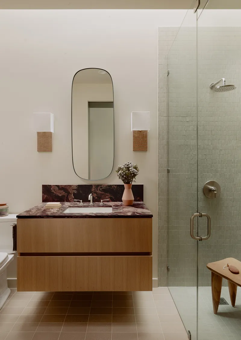 Floating oak vanity with marbled countertop beside a glass-enclosed shower with pale green mosaic tile