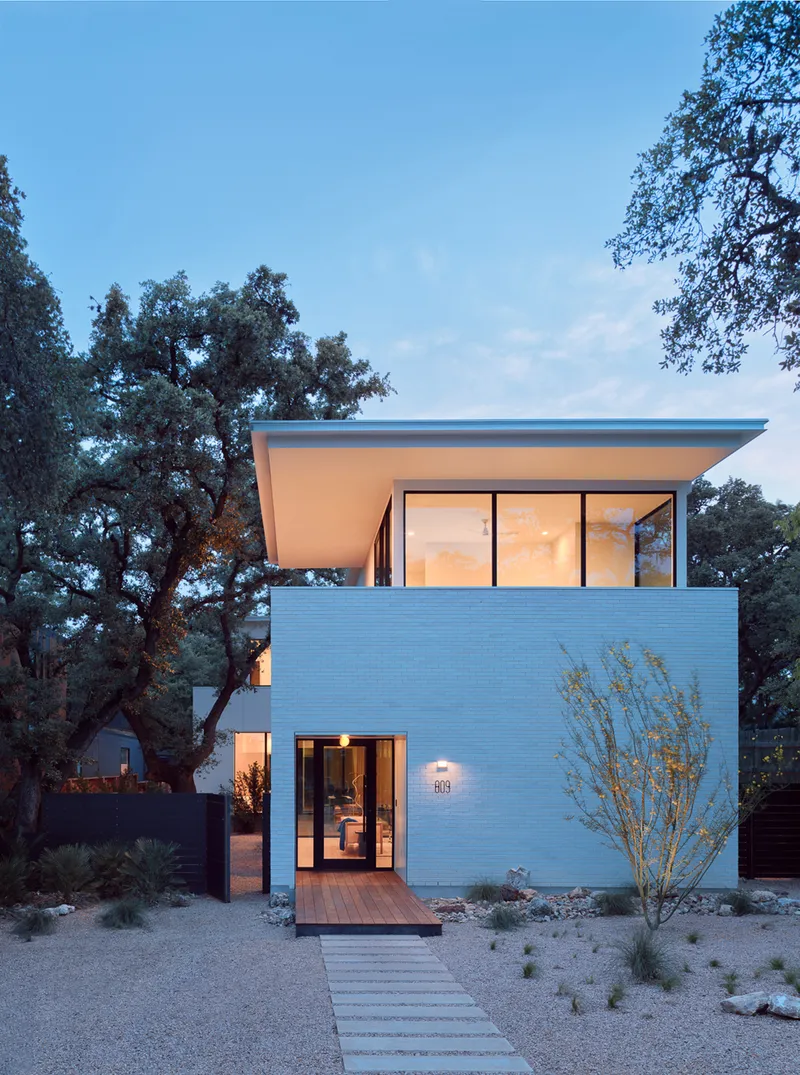 Two-story white brick volume with floor-to-ceiling glass doors illuminated at dusk beside an old oak tree