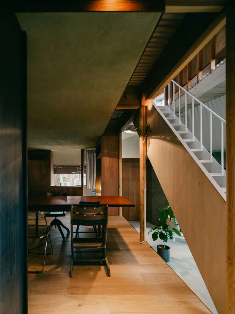 Dining area with timber table beneath a white-painted staircase and exposed plywood ceiling