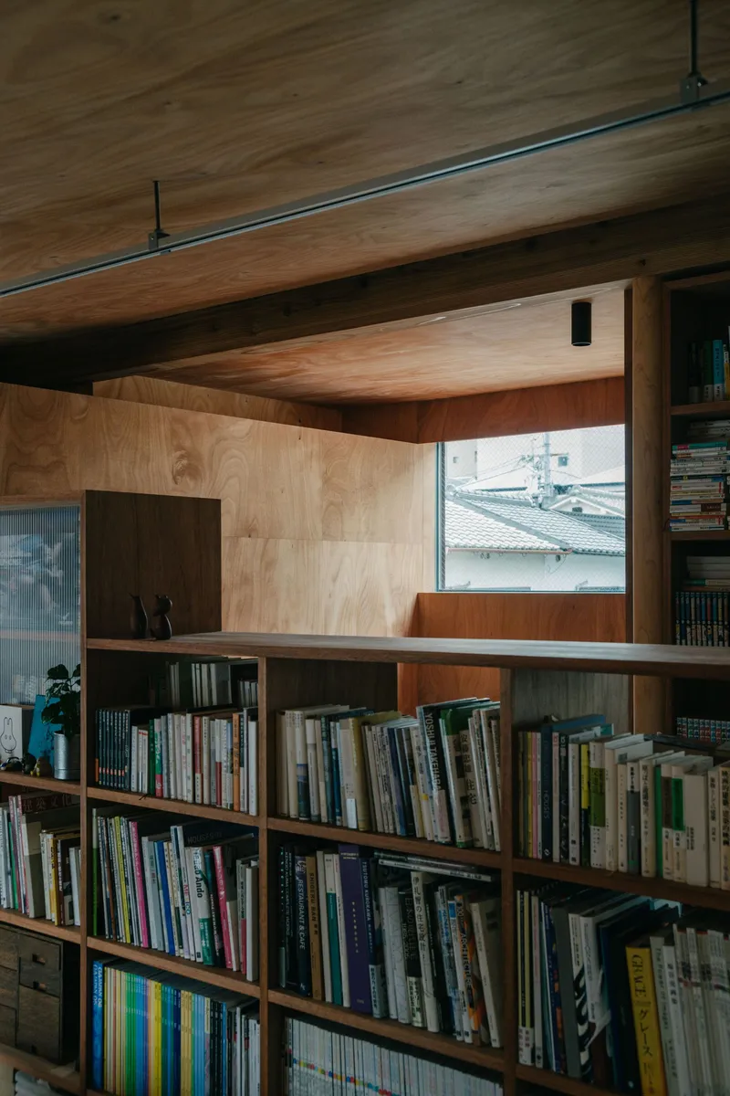 Built-in plywood bookshelf under timber ceiling with horizontal window framing tiled rooftops